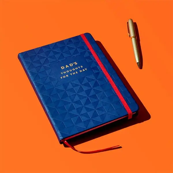 A blue textured notebook titled Dad’s Thoughts for the Day with a red elastic strap, shown with a matching pen on a bright orange background.